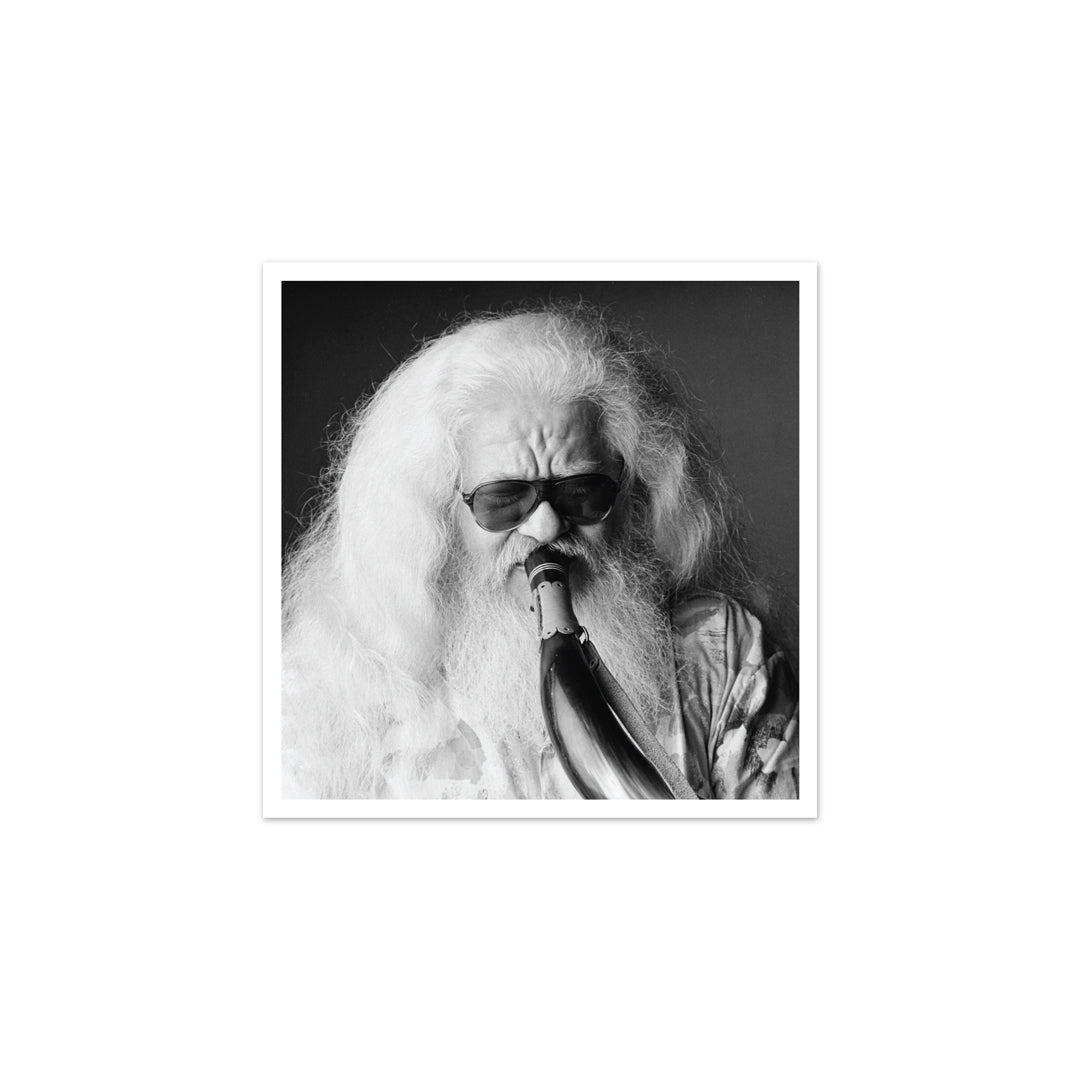 Fotografia artística em preto e branco com moldura branca, focando Hermeto Pascoal em grande plano de óculos escuros e barba longa branca, tocando um instrumento de sopro feito de chifre.