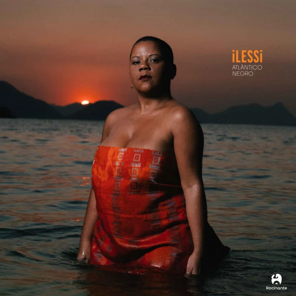 Capa do LP Atlântico Negro de ILESSI com artista em pé no mar ao entardecer, vestindo roupa vermelha e paisagem costeira ao fundo durante o pôr do sol (disco de vinil).