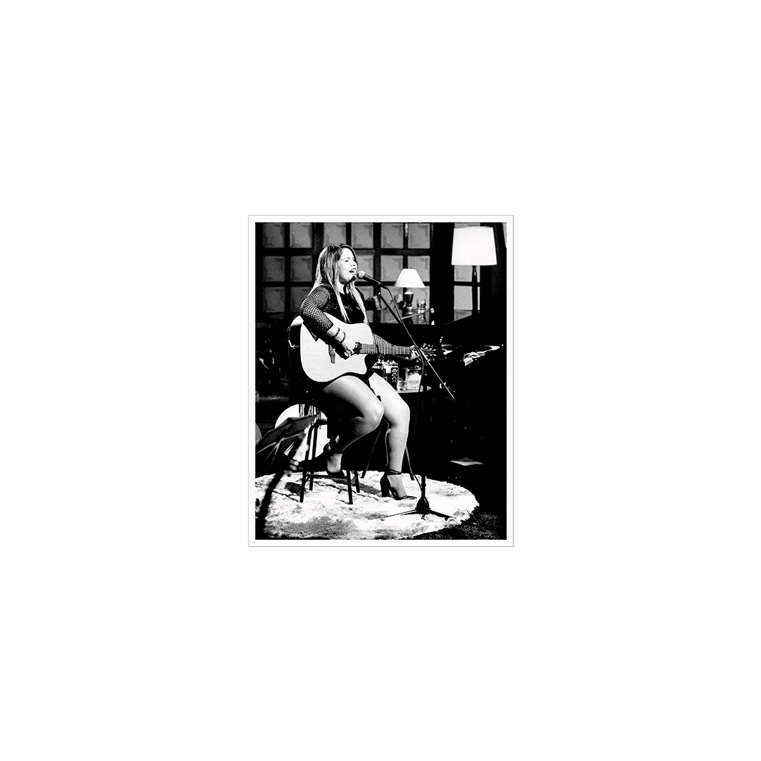Cartão fotográfico em preto e branco incluído na edição em vinil de Ao Vivo, de Marília Mendonça, mostrando a cantora sentada em um banco durante a gravação, cantando ao microfone enquanto toca violão. Ao fundo aparecem luminárias, piano e elementos do estúdio onde a apresentação foi registrada.